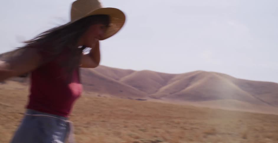 A joyful woman in a straw hat runs and dances through a vast golden field with rolling hills on a sunny day