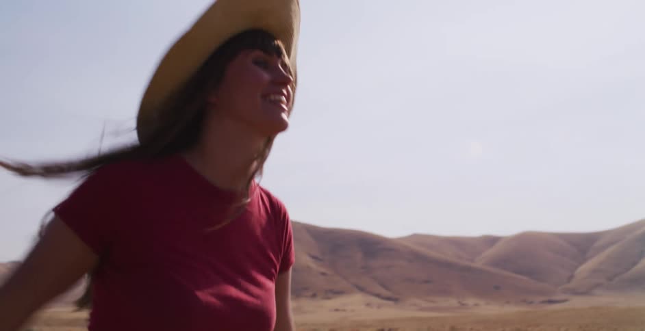 A carefree woman in a straw hat smiles as she runs in slow motion through a vast, golden field with arid hills behind her