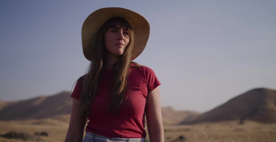 Close-up of a thoughtful woman in a straw hat standing in a dry, sunlit field with hills in the background
