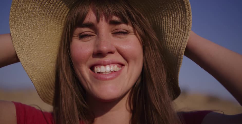 A beautiful young woman in a straw hat looks into the camera and breaks into a genuine, happy smile in slow motion