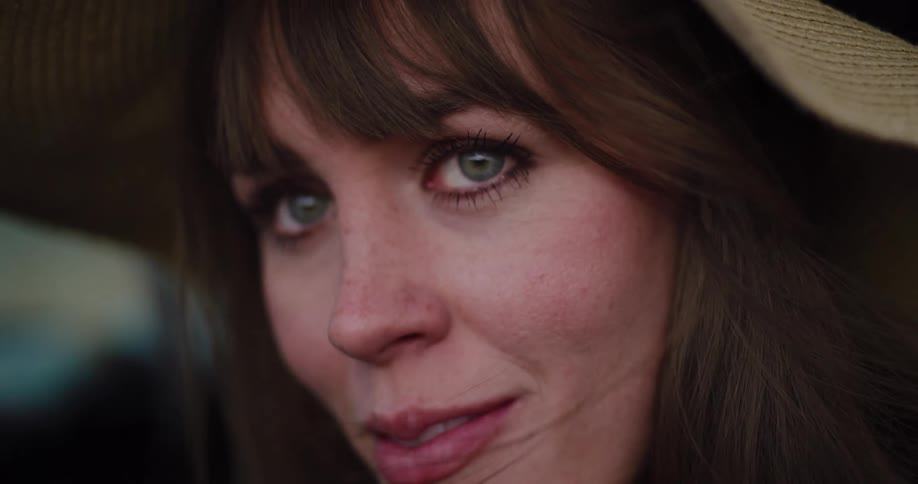 A serene woman with green eyes wearing a hat looks into the camera as wind blows through her hair in slow motion