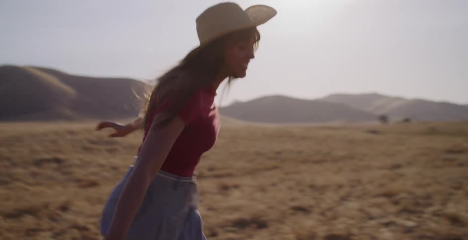 A carefree woman in a red shirt and cowboy hat joyfully runs through a sunny, golden landscape in slow motion