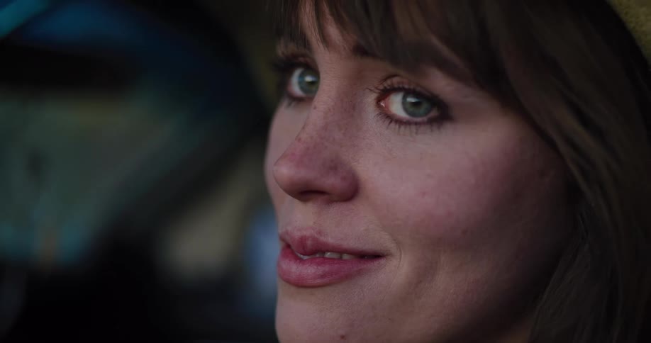 A beautiful woman with green eyes and a wide-brimmed hat smiles contemplatively in an intimate, slow-motion close-up as she sits in a vehicle
