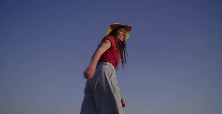 A joyful young woman in a straw hat smiles and spins in slow motion against a brilliant blue sky