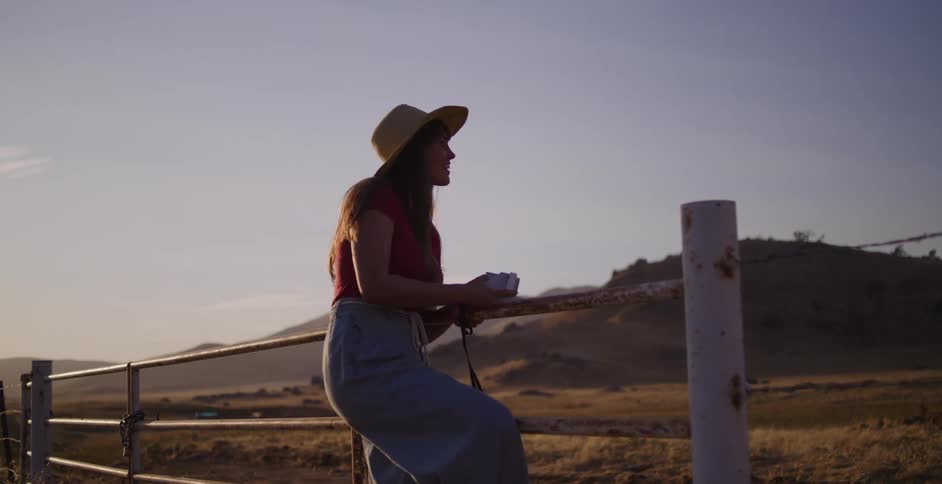 A young woman on a road trip climbs a rural fence and takes a picture of the landscape with an instant camera during a beautiful golden hour sunset