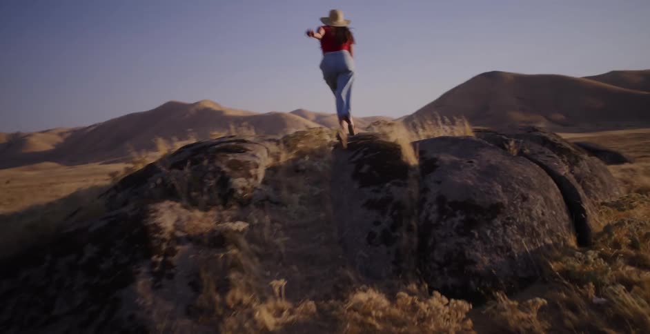 A free-spirited woman in a straw hat runs up a rocky hill in a golden field and spreads her arms in a moment of pure freedom