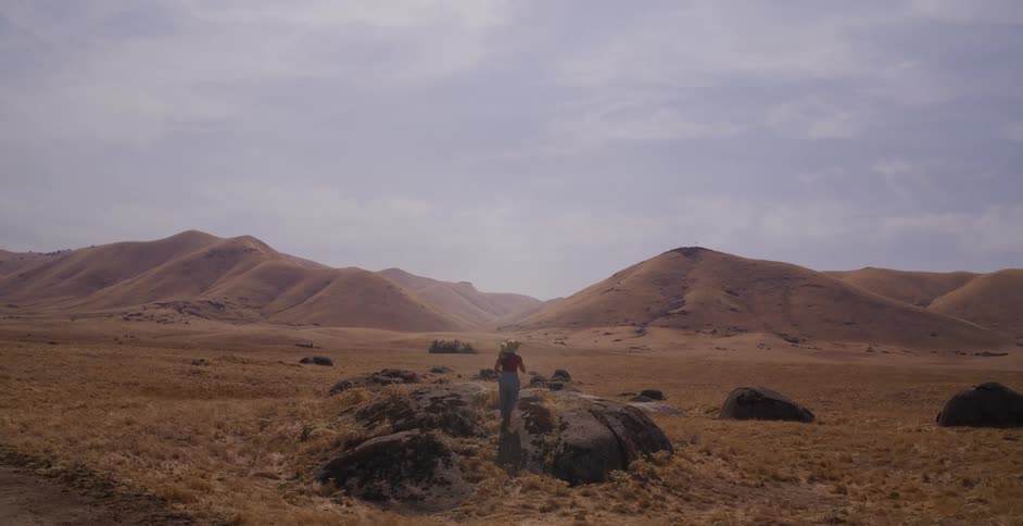 A lone woman wearing a straw hat explores a vast and arid landscape, climbing a large boulder to survey the rolling golden hills