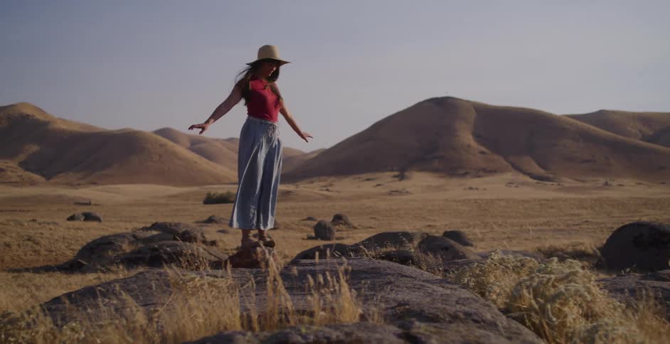 Slow motion shot of a woman in a sun hat and long pants balancing on rocks in a vast, dry, hilly landscape