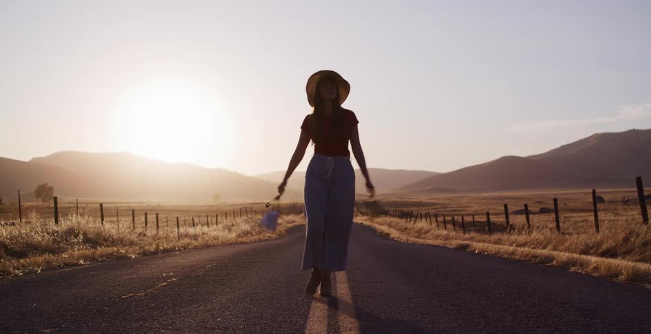 Slow motion shot of a woman in a wide-brimmed hat walking towards the camera on a rural road at sunset