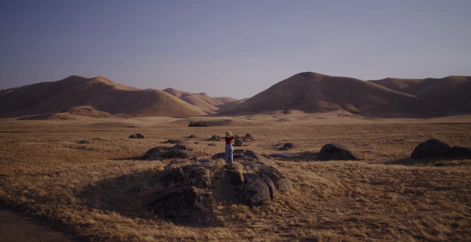 A woman in a straw hat runs through a golden, hilly landscape and triumphantly raises her arms after climbing a large rock