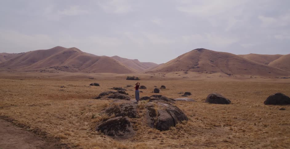 A woman in a straw hat runs across a vast, golden, rocky landscape and stands on a boulder with her arms outstretched in freedom