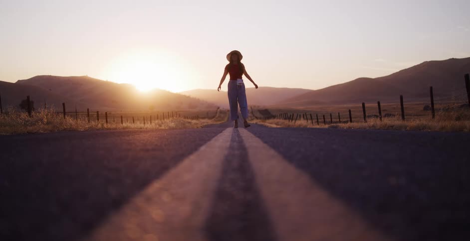 A free-spirited woman walks down the center of a deserted country road at sunset, silhouetted by the golden light