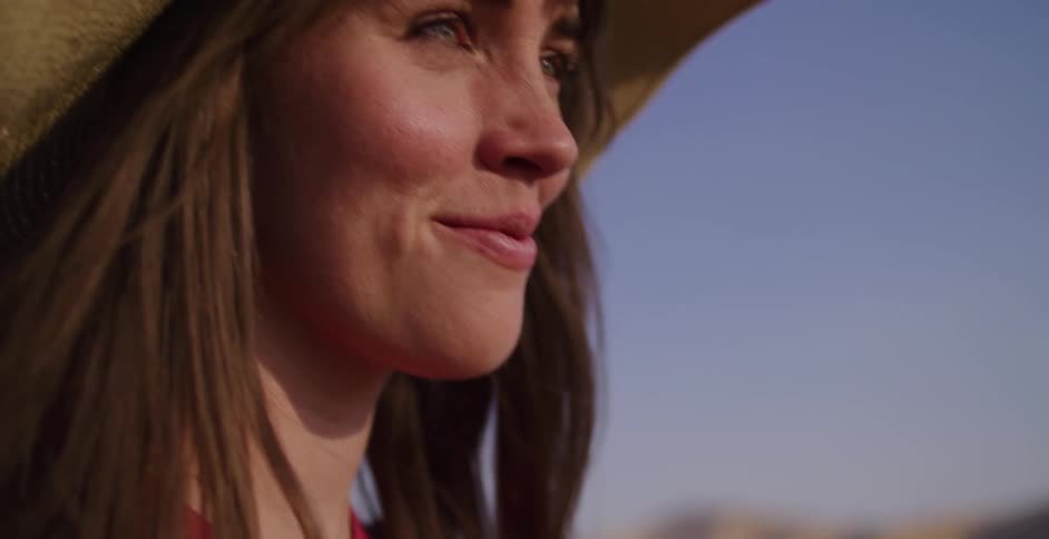 Close-up of a woman in a straw hat as she looks out and smiles, her hair blowing in the wind during golden hour