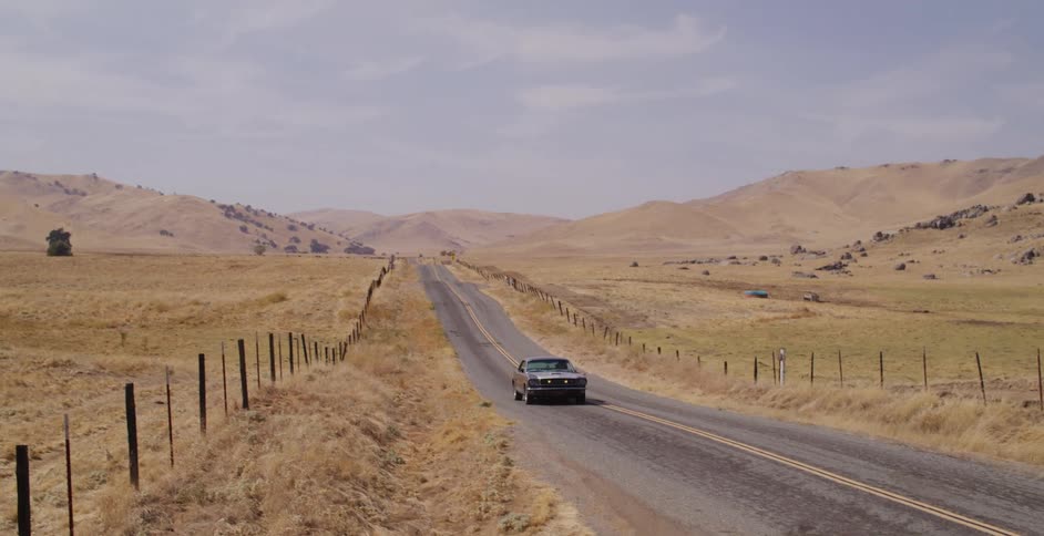 A silver classic muscle car drives down a deserted two-lane highway through dry, golden hills