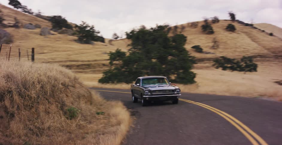 A classic black Mustang cruises along a scenic, winding road in the rural countryside