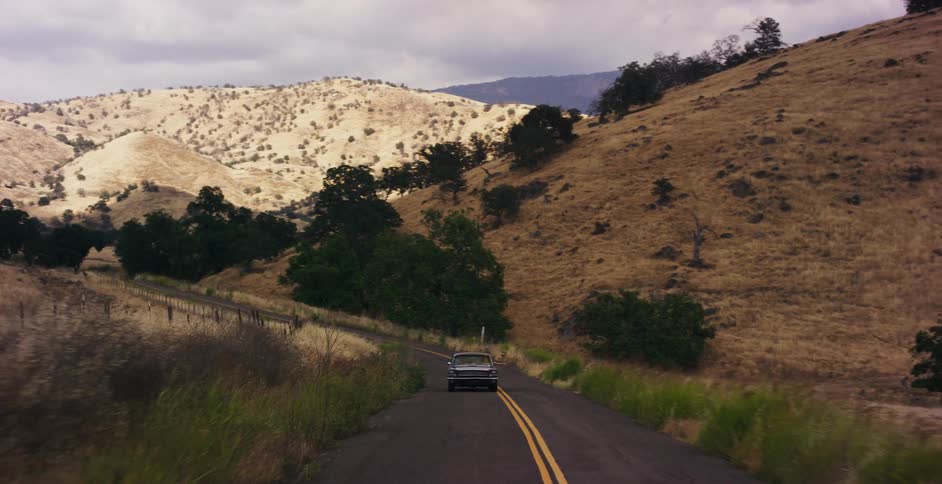 A classic car cruises down a lonely country road through golden rolling hills under an overcast sky