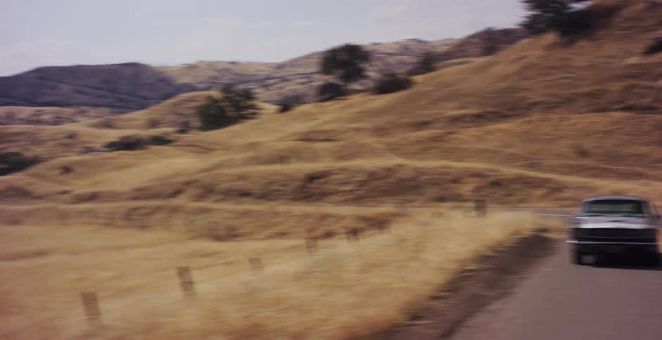 A classic silver Mustang drives on a winding road through dry, golden hills before the camera pans to the vast landscape