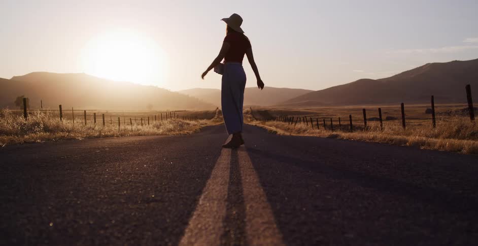 A silhouetted woman walks freely down an empty country road at sunset before stopping to take a photograph
