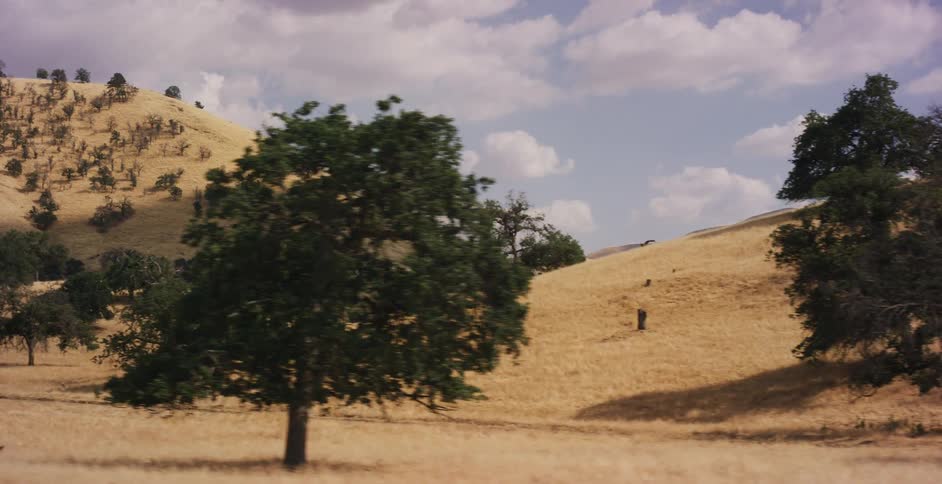 A slow pan across a landscape of dry, golden hills dotted with green oak trees under a partly cloudy sky