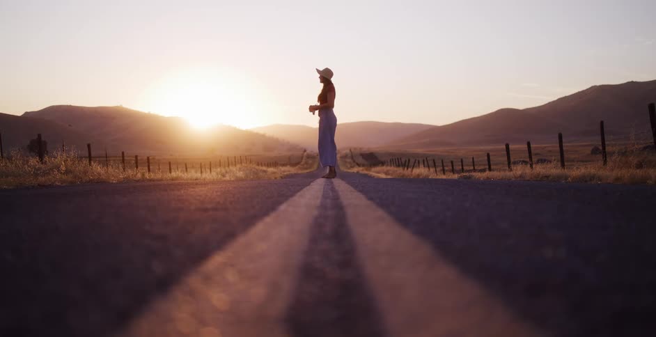 A carefree woman walks and balances on the yellow lines of a deserted country road during a beautiful golden hour sunset