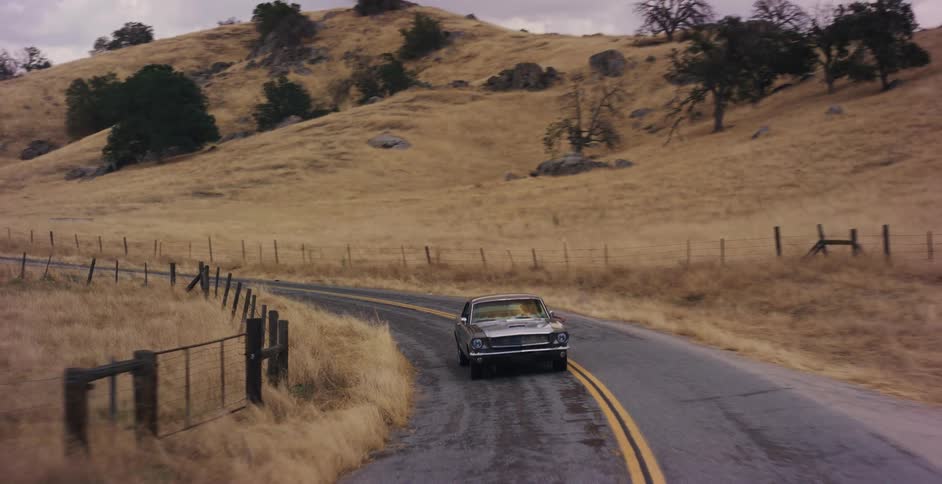 A vintage black sports car cruises down a winding rural road through dry, golden hills on an overcast day