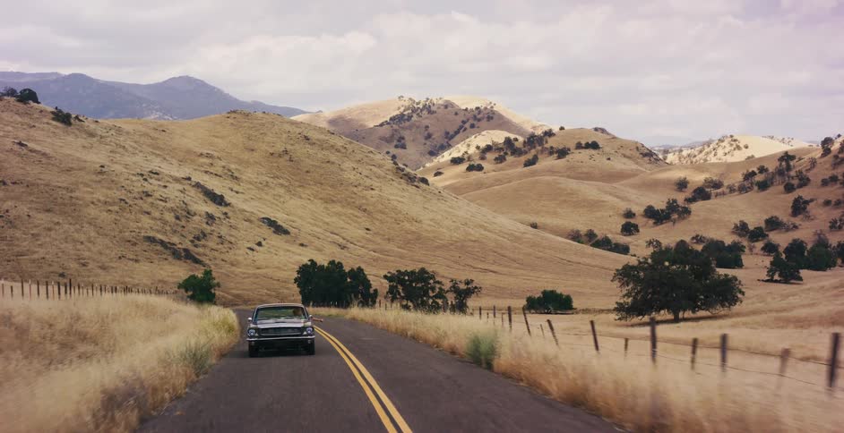 A classic black American muscle car cruises down a scenic country road through the dry golden hills of California