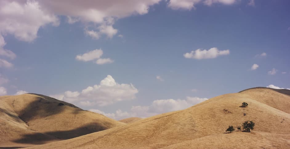 A slow aerial drone flight ascending over vast, dry golden hills under a beautiful blue sky with scattered clouds