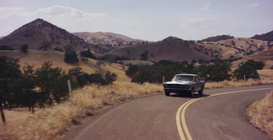 A vintage silver muscle car is parked on a winding country road amidst rolling golden hills on a sunny day