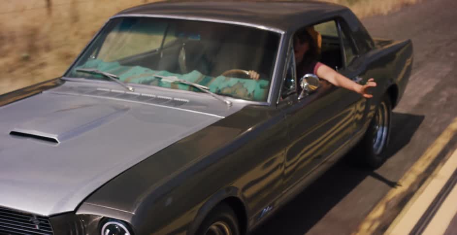 A carefree woman in a hat drives her classic Ford Mustang down a winding country road with her arm out the window on a sunny day