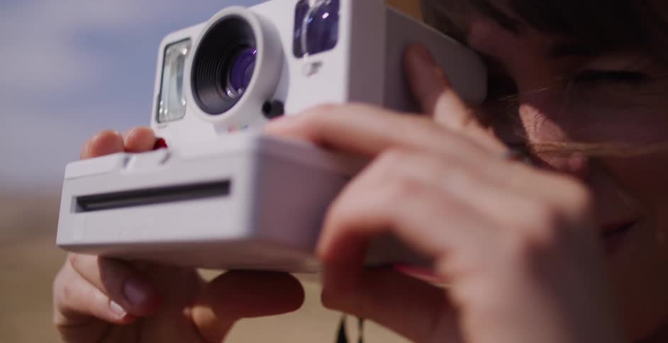A close-up of a smiling woman in a straw hat bringing a retro instant camera to her eye to take a photograph on a sunny day