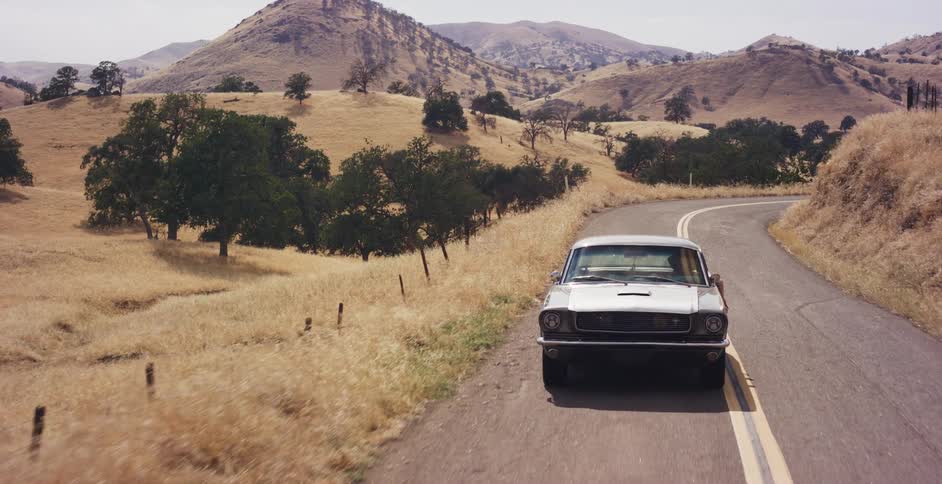 A classic silver Ford Mustang drives along a winding country road through dry, golden hills on a sunny day