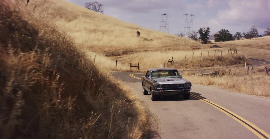 A classic silver Ford Mustang drives slowly down a winding country road surrounded by golden hills