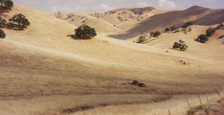 A vintage Ford Mustang drives on a winding road through a dry, golden rural landscape in slow motion