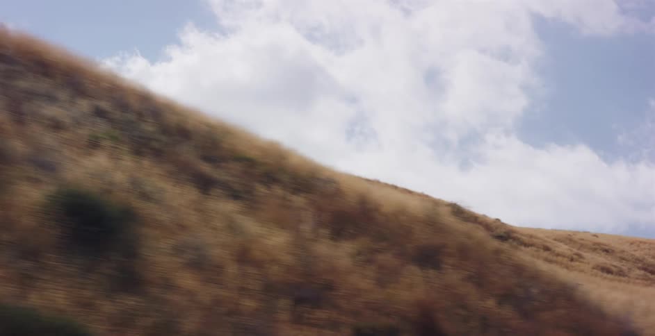 A tracking shot captures the motion blur of a dry, golden grass hillside against a partly cloudy sky