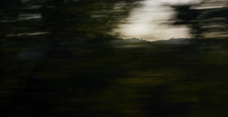 A dreamy, motion-blurred view of rolling green hills and hazy mountains speeds by from a moving vehicle's window
