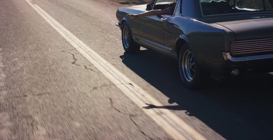 A cinematic slow-motion shot follows a classic muscle car as it cruises down a sun-drenched road, with a hand hanging freely out the window