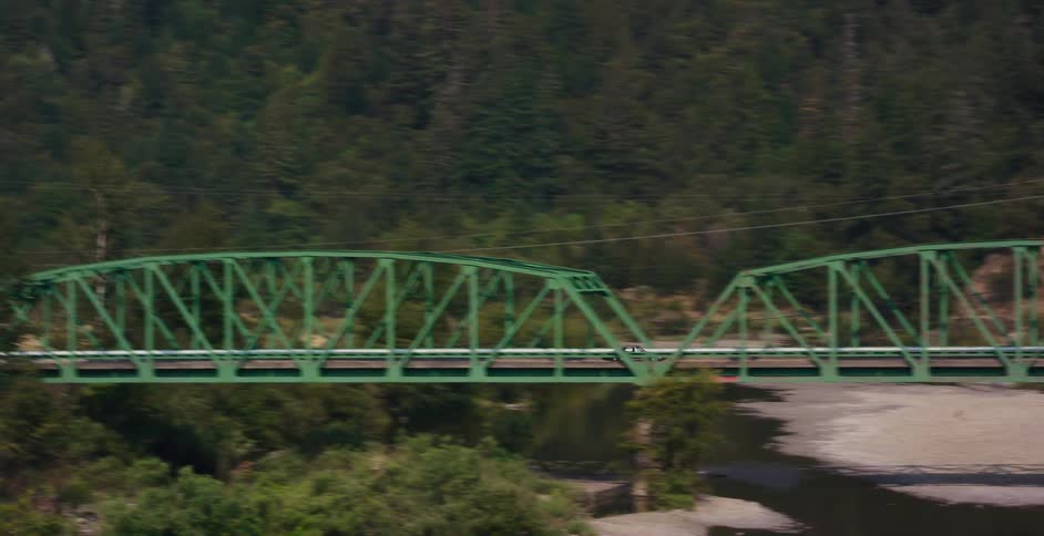 A scenic tracking shot reveals a green steel truss bridge spanning a river in a lush, forested valley