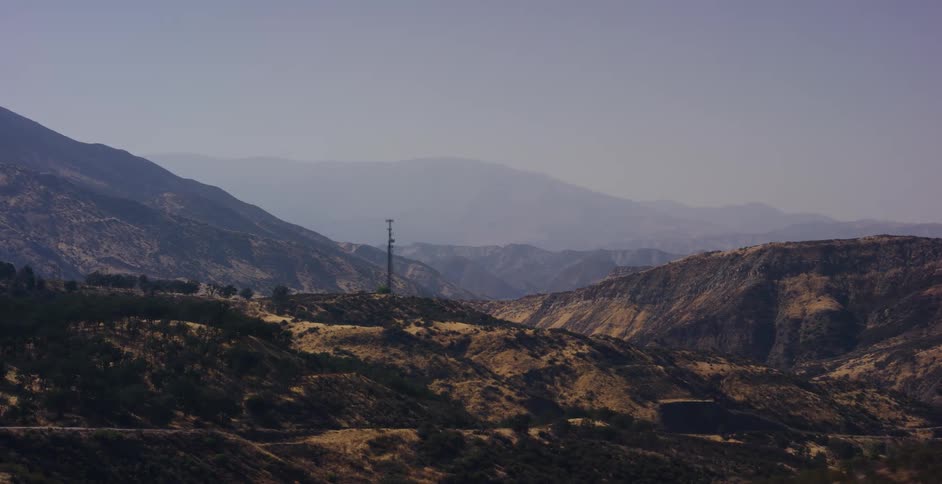 A scenic driving shot reveals a vast, arid mountain range with a lone communication tower under a hazy sky