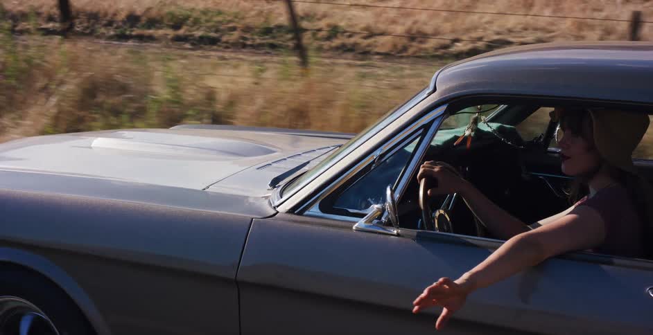A woman in a sun hat enjoys a leisurely drive in a vintage Ford Mustang through the sunny, golden hills of the countryside