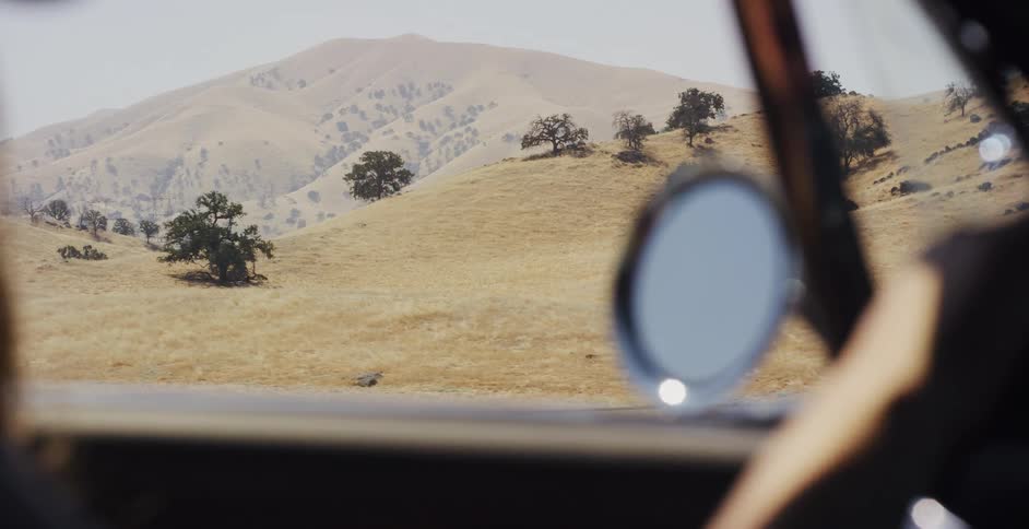 A scenic view of golden, rolling hills and oak trees passes by through the window of a car on a road trip