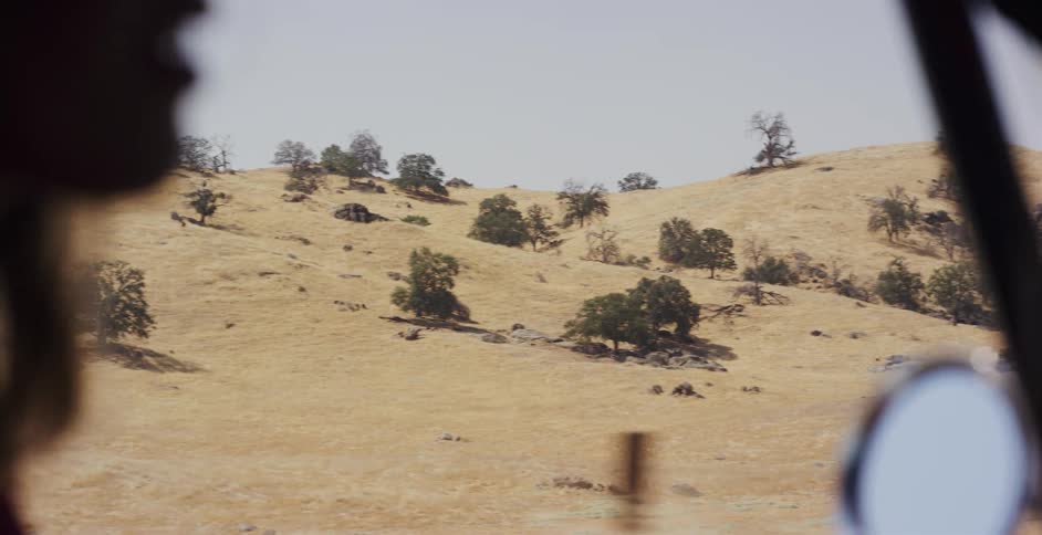 A slow-motion tracking shot from inside a vehicle reveals a sun-drenched, golden hillside dotted with oak trees