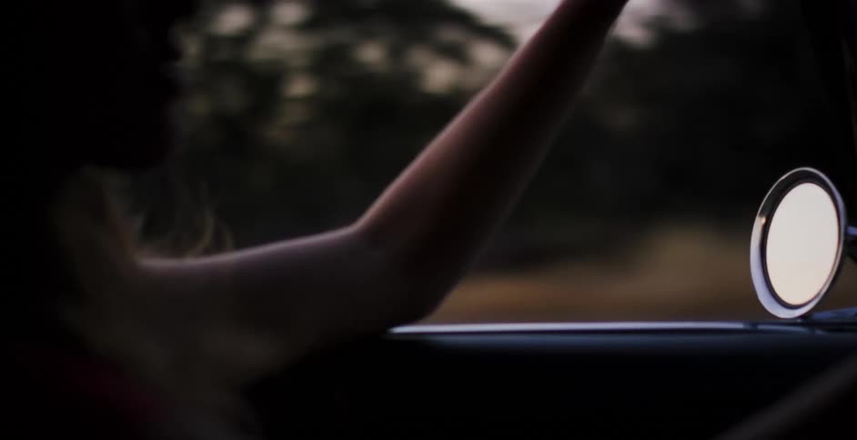 A silhouetted woman enjoys a scenic drive through the countryside from the passenger seat of a car at sunset