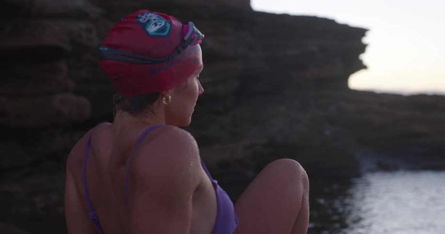 A female swimmer in a purple swimsuit and red swim cap sits on a rocky coastline, gazing out at the ocean at sunrise