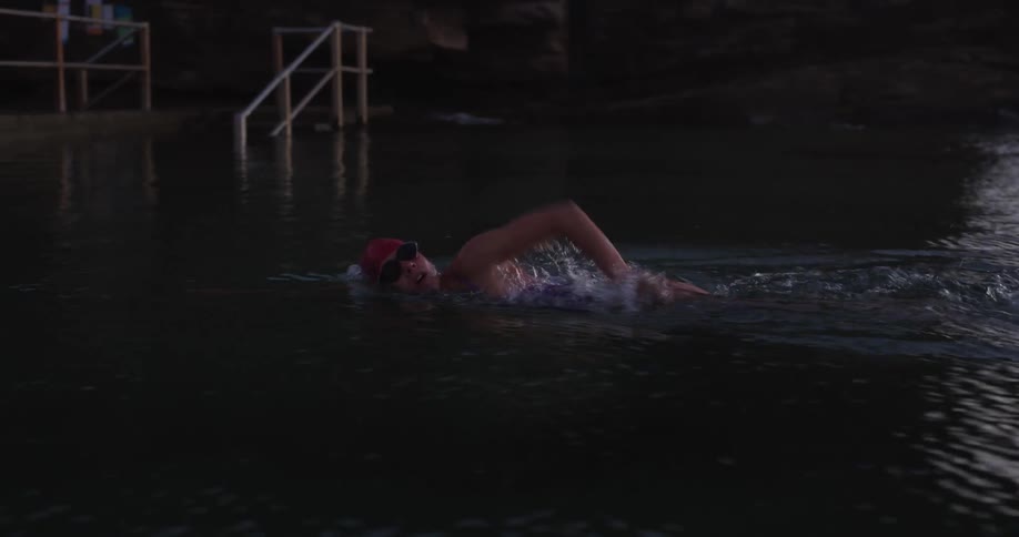 A determined female swimmer practices her freestyle stroke in a dark ocean pool at dusk