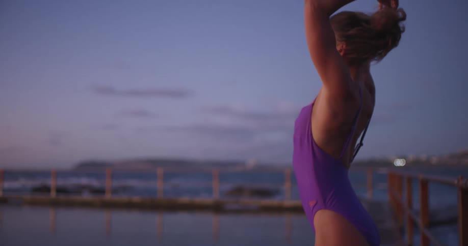 A woman in a purple swimsuit ties her hair and prepares her swimming cap by an ocean pool at dusk in slow motion