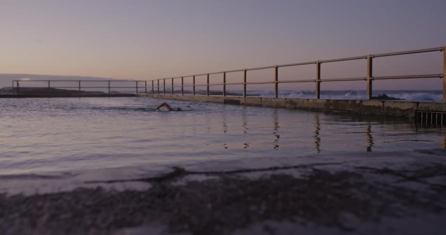 A swimmer gracefully moves through a tranquil ocean rock pool in slow motion at sunrise, with waves crashing in the background