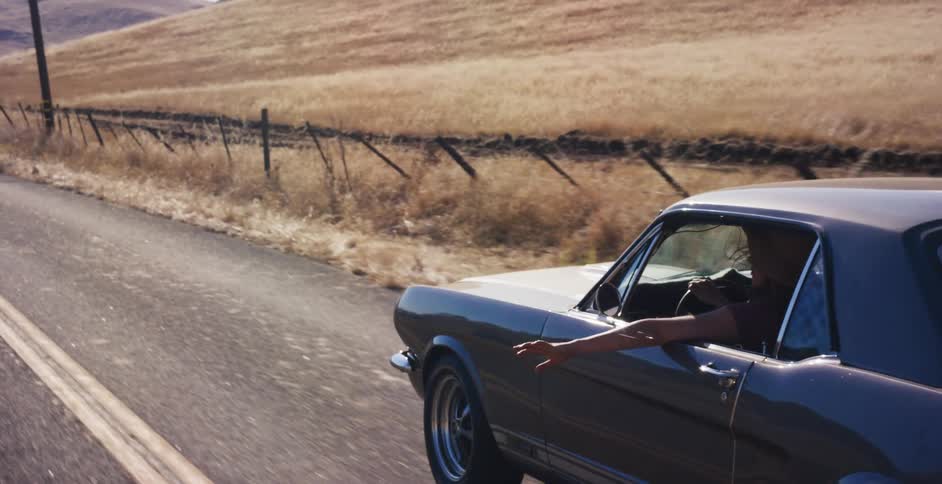 A woman in a classic Ford Mustang enjoys a scenic drive down a country road with her arm out the window on a sunny day