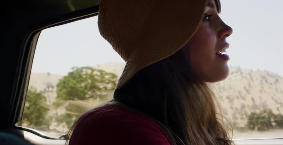 A young woman wearing a yellow hat enjoys the view from the passenger seat of a car on a sunny day road trip