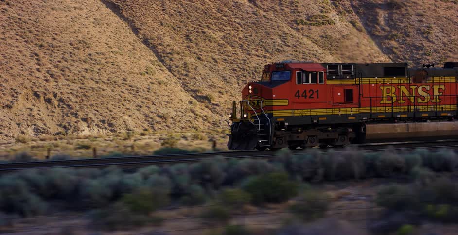 A BNSF freight train locomotive travels along tracks through a dry, hilly desert landscape at golden hour