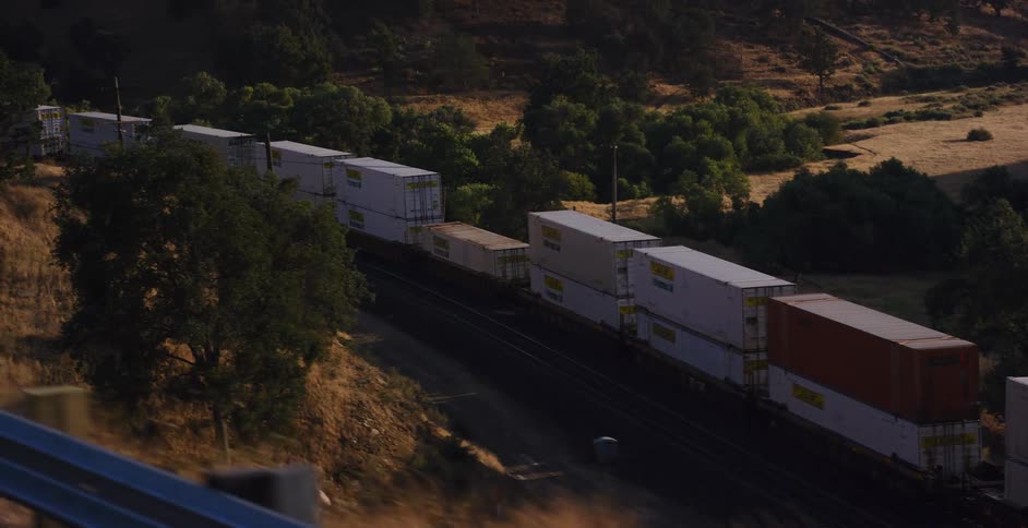 High angle shot of a freight train carrying shipping containers as it moves through a green and golden hilly landscape